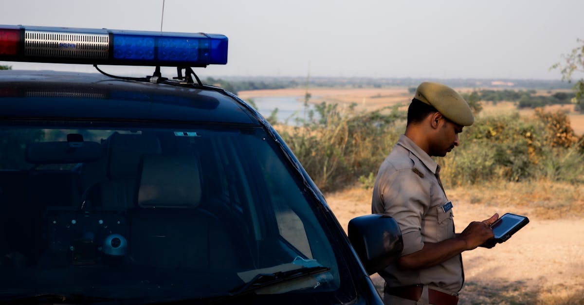 A police officer using a tablet beside a patrol car in a rural outdoor setting.